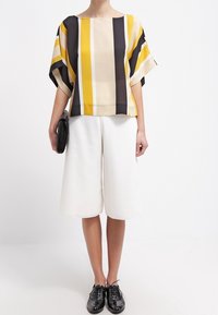 W paski bluzka w kolorach żółtym, czarnym i beżowym, w połączeniu z białymi kulotami. Modelka trzyma czarną torebkę kopertową, ma na sobie czarne buty. Prosty design, swobodny fason.
