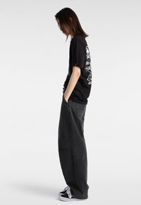 Camiseta negra con estampado gráfico en la espalda, combinada con jeans sueltos de color gris oscuro. El modelo lleva zapatillas negras y blancas, con las manos en los bolsillos.