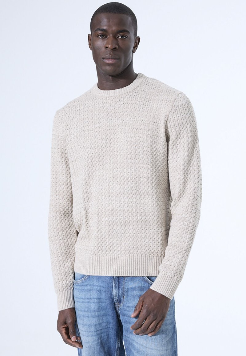 Beige gestrickter Pullover mit strukturiertem Muster und gerippten Bündchen. Kombiniert mit blauen Jeans, vor einem schlichten weißen Hintergrund präsentiert.