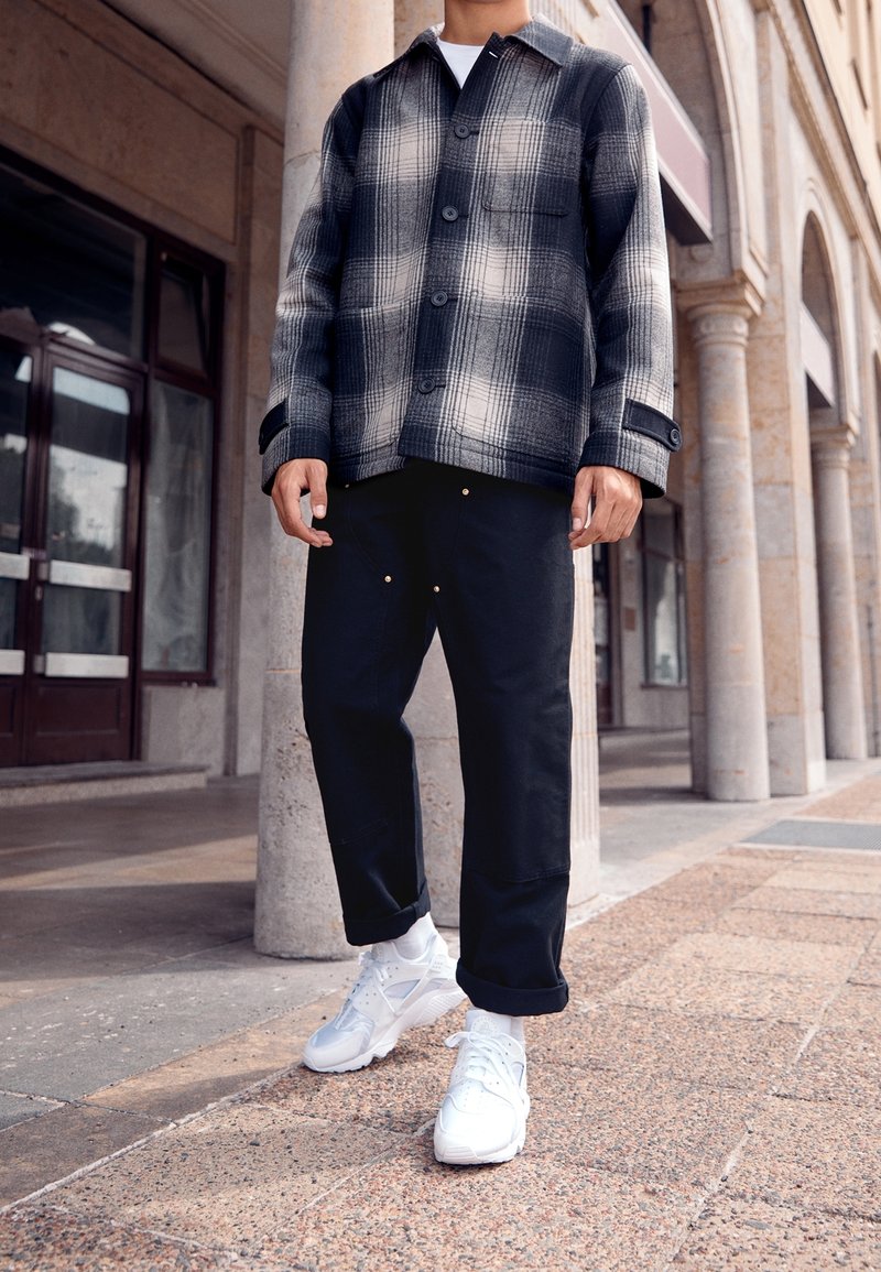 Giacca a quadri in bianco e nero con chiusura a bottoni, pantaloni neri e sneakers bianche. Tessuto liscio e vestibilità casual; i pantaloni hanno dettagli dorati.