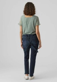 Hellgrüne, gemusterte Bluse mit Rüschenärmeln, kombiniert mit eng geschnittenen, dunkelblauen Jeans aus Denim, die hinten mit überkreuzten Trägern ausgestattet sind.