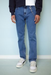 Person med blå jeans med en kedja fäst vid sig och vita sneakers som står mot en ljusblå vägg och golv.