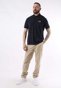 Camisa polo azul marino con collar y mangas cortas, pantalones beige claros y zapatillas blancas; tiene un logo visible en el pecho.