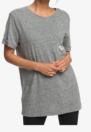 Woman wearing a loose grey t-shirt with rolled-up sleeves and a small logo on the left chest pocket, hands clasped in front.