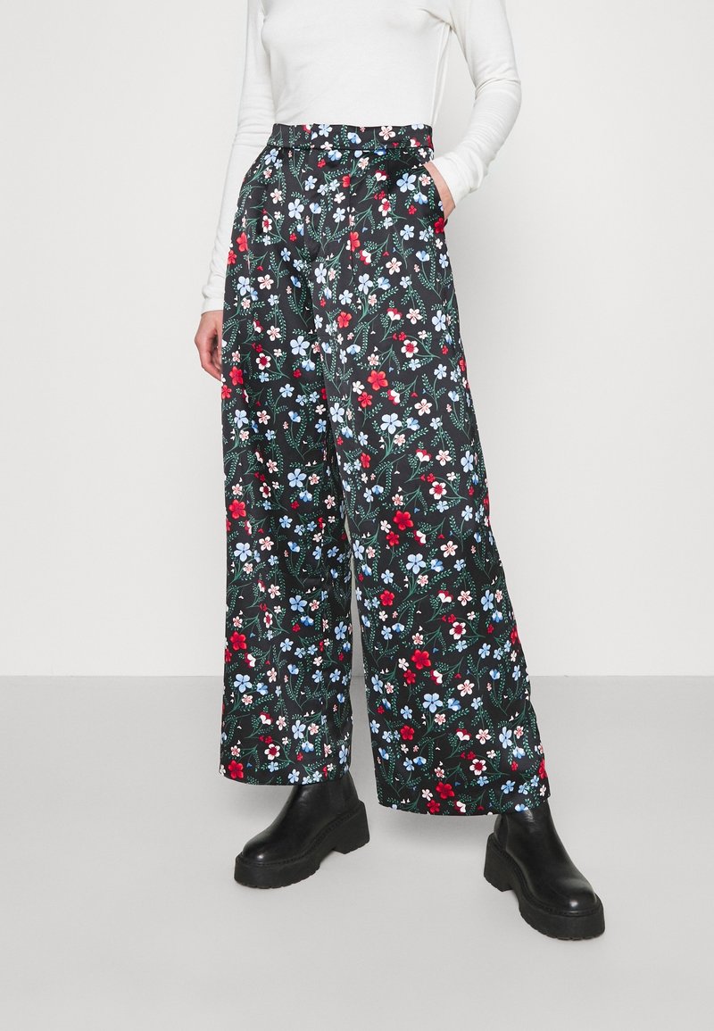 Personne portant un pantalon noir à taille haute et jambes larges avec un imprimé floral rouge, bleu et blanc, un haut blanc à manches longues et des bottes à plateforme noires.