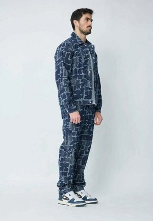 Veste en denim et pantalon bleu foncé avec des motifs géométriques. Accompagnés de baskets blanches. Design slim fit, tissu texturé.