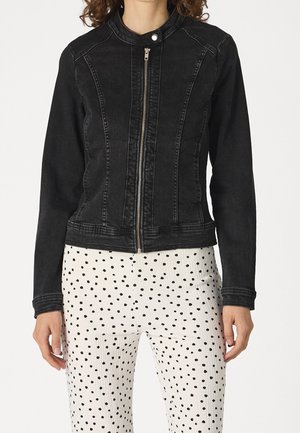 Femme portant une veste en jean noire zippée et un pantalon blanc à pois noirs, debout devant un fond blanc uni.