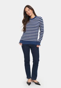 Jūrinės ir grietinėlės spalvų dryžuotas megztinis su ilgomis rankovėmis, raišteliais ant rankovių ir apatinės dalies, derinamas su tamsiomis džinsomis ir smailiais juodais bateliais.