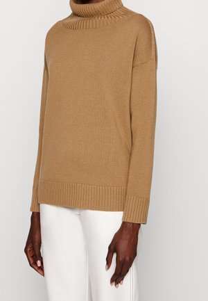 Persoon in een trui met hoge hals in kamelkleur, gecombineerd met witte broek, staand tegen een effen lichte achtergrond.