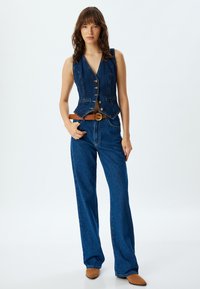 Denimli yelek, uyumlu bir tasarıma sahip, beş düğme ve iki ön cep ile donatılmış; geniş paçalı denim pantolon ve kahverengi bir kemer ile kombinlenmiştir.