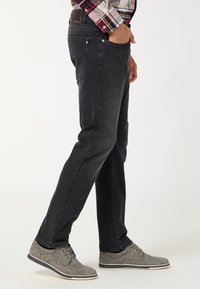 Jeans in denim nero con vestibilità slim, dotati di cinque tasche e una leggera texture sbiadita. Abbinati a scarpe stringate grigio chiaro.