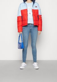 Persona che indossa una giacca puffer color block rossa e azzurro chiaro, una felpa grafica azzurro chiaro, jeans skinny azzurro chiaro, sneakers bianche e tiene in mano una borsa blu.