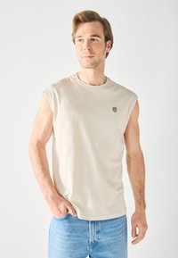 Cream sleeveless t-shirt in soft fabric with a round neck, featuring a small green logo patch on the left chest area. Paired with blue jeans.