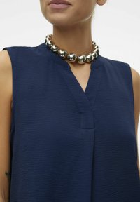 Débardeur sans manches navy avec un tissu texturé et un décolleté en V, associé à un collier en perles métalliques brillantes.