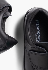 Chaussures en cuir noir à bout arrondi et bande velcro. L'intérieur présente "Pisanias, Hecho en España" embossé en argent. Texture lisse.