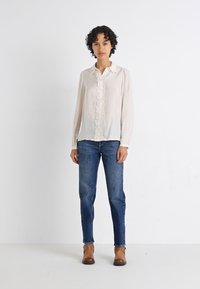 Blusa de crema clara con frente Volante, mangas largas y textura suave, combinada con pantalones vaqueros azul denim y botas de tobillo marrones.