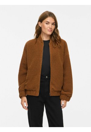 Femme aux longs cheveux bruns portant une veste teddy marron par-dessus une chemise noire et un pantalon noir, debout devant un fond blanc uni.