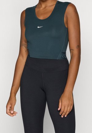 Žena nosící tmavě zelený krátký crop top Nike bez rukávů a černé legíny s vysokým pasem, ukazující tetování na obou pažích.