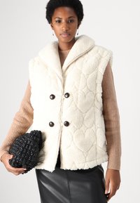 Krämig shearling-väst med en stor krage, kviltad hjärt-mönster och mörka knappar. Kombinerad med en ribbad brun tröja och en texturerad svart väska.