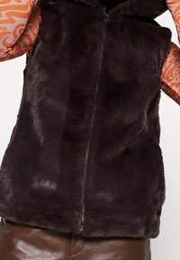 Dunkelbrauner Kunstfell-Weste mit Reißverschluss und Kapuze. Weicher Stoff, ärmelloses Design, kombiniert mit einem orangefarbenen, gemusterten Langarmshirt.