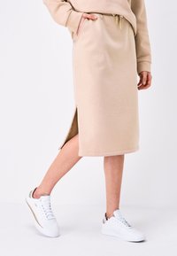 Jupe midi beige en tissu doux, avec une fente latérale, taille élastique avec cordon de serrage, portée avec des baskets blanches. Design minimaliste.