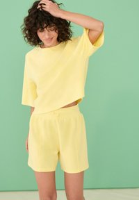 Ensemble de vêtements de détente en coton jaune comprenant un haut oversized à manches courtes et un short à taille élastique, sur fond vert menthe.