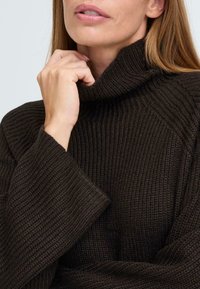 Dunkelbrauner Strickpullover mit hohem Rollkragen und weiten Ärmeln, der ein strukturiertes Rippmuster aufweist. Nahaufnahme des Stoffdetails.