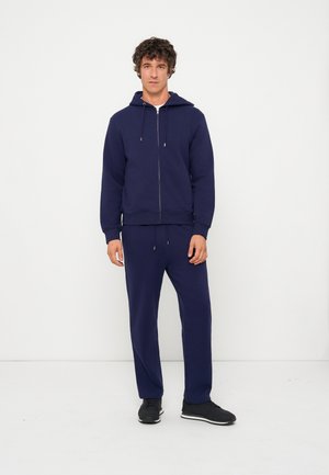 Giovane uomo in piedi che indossa una felpa con cappuccio blu navy con zip e pantaloni della tuta coordinati con sneakers nere su uno sfondo bianco semplice.