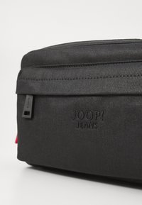 Borsa in tessuto nero con texture liscia, dotata di chiusura a zip e logo in rilievo "JOOP! JEANS", insieme a una linguetta di accento rossa.