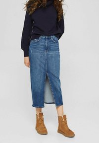 Jupe midi en denim avec une fente sur le devant, présentant un délavage clair, deux poches et une silhouette ajustée, associée à des bottes à lacets beiges.