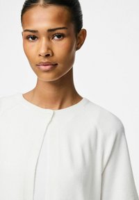 Balta poguņu kardigāna jaka ar mīkstu tekstūru, raglu piedurknēm un apaļu kakla izgriezumu. Minimālisma dizains bez redzamām rakstu vai akcentu detaļām.