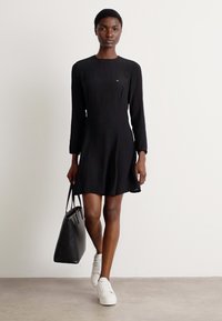 Melnas garām piedurknēm kleita ar pieguļošu bodi un izplestu svārku, apvienota ar melnu somu un baltām sporta apaviem. Vienkāršs dizains, gluda auduma.