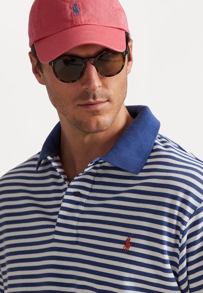 Vīrietis valkā brūnus bruņurupača raksta saulesbrilles, sarkanu beisbola cepuri un zilas ar baltām svītrām polo krekls ar zilu apkakli.