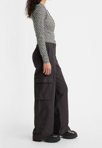 Vrouw in zwarte wijde cargobroek, zwarte chunky schoenen en een zwart-witte bloemen lange mouw top, staande in profiel op een witte achtergrond.