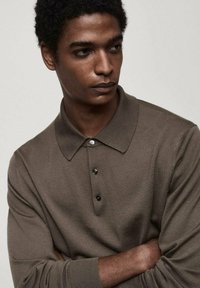 Polo en tricot marron avec col, trois boutons noirs et poignets côtelés. Le modèle pose les bras croisés devant un fond neutre.