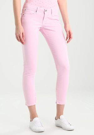 Vrouw draagt lichtroze skinny jeans en witte sneakers, staand met de armen ontspannen langs het lichaam tegen een effen achtergrond.