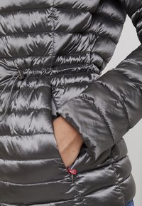 Graue Pufferjacke mit glänzender Oberfläche, ausgestattet mit einem Kordelzug in der Taille, Seitentaschen und einem gesteppte Muster zur Isolierung.