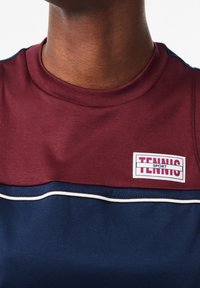 Vinröd och marinblå sportskjorta med rundad hals, horisontell färgblockdesign, som har en logotyppatch med texten "TENNIS SPORT" och vita kantband.