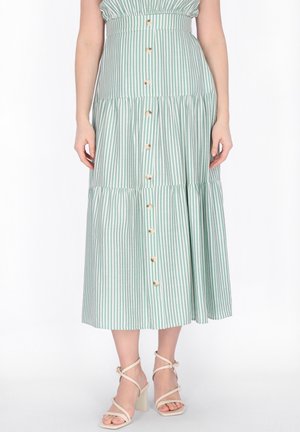 Frau trägt einen grün-weiß vertikal gestreiften, knielangen Rock mit Knöpfen und beige Riemchen-Sandaletten mit hohen Absätzen.