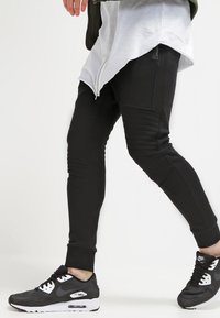 Černé slim-fit tepláky s žebrovanými detaily, kombinované s černými teniskami Nike se bílou podrážkou a logem. Pod tím bílá vrstvená košile.