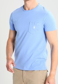 Světle modré bavlněné tričko s krátkými rukávy, kulatým výstřihem a kapsou na hrudi, která má malé vyšívané logo.