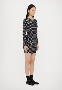 Long-sleeve black dress with white polka dots, fitted design, and rounded neckline. Model wearing black socks and flats.