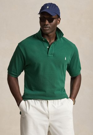 Férfi sötétzöld galléros pólóban, fehér nadrágban, tengerészkék sapkában, napszemüvegben és karórával, zsebében tartva kezét.