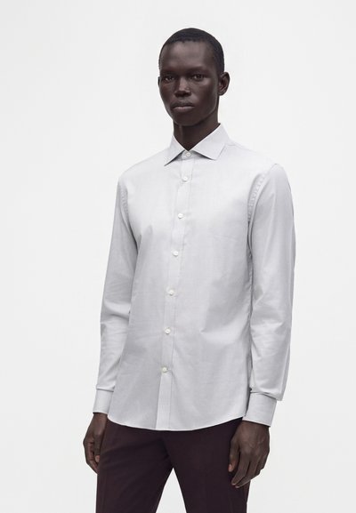 Homme portant une chemise blanche à manches longues avec col pointu et pantalon foncé, debout devant un fond blanc uni.