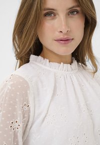 Blouse blanche en tissu texturé, ornée de broderies et de motifs ajourés. Encolure à volants et manches transparentes à motifs.