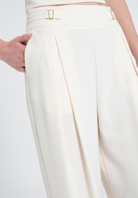 Pantaloni bianchi a pieghe realizzati in tessuto morbido, dotati di una vita piatta con una fibbia dorata e tasche laterali.