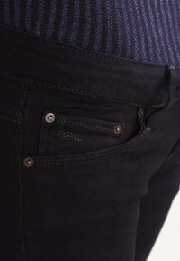 Pantalones vaqueros de mezclilla negra con un bolsillo frontal que presenta herrajes plateados y sutiles detalles de costura. La tela parece suave y ajustada.