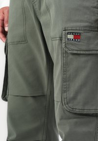 Close-up of olive green cargo pants with a flap pocket and Tommy Jeans logo patch near the thigh.