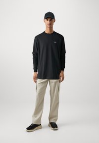 Svart langermet t-skjorte med en liten logo, kombinert med beige bukser og svarte joggesko med tykk såle. En svart cap fullfører antrekket.