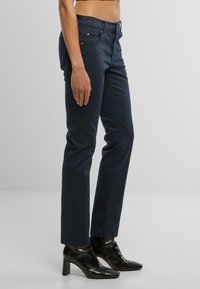 Jeans de mezclilla azul marino ajustados, con cremallera en la bragueta, cinco bolsillos y sutiles acentos metálicos. Llevados con botas de tacón alto negras hasta el tobillo.